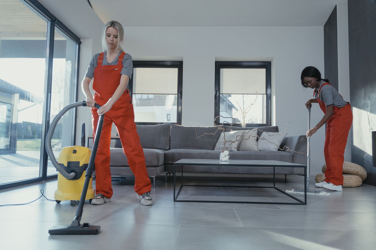 services-02 Two women in red overalls vacuuming and sweeping a modern living room.